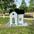 White bird feeder shaped like a house with a black roof, hanging outdoors.