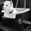 Small white dog sitting in a black pet car seat with a fluffy white blanket inside a vehicle.